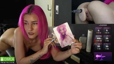 Media: A video shows a topless woman with pink hair, holding a pink phone displaying a woman's naked body. The background features a dark room with a bed and a screen displaying stats.