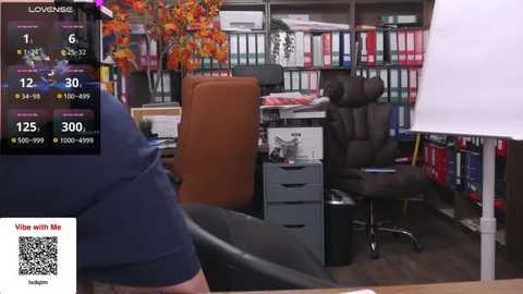 Media: Video of a cluttered office with a large wooden desk, black office chair, and colorful bookshelves. A person with a dark skin tone, wearing a black shirt, is seen from the back.