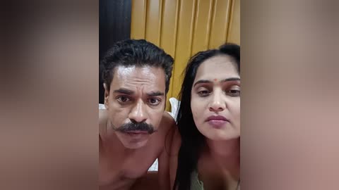 Media: Video of a man with a mustache and woman with dark hair, both topless, standing closely together in front of yellow curtains, giving a suggestive pose.