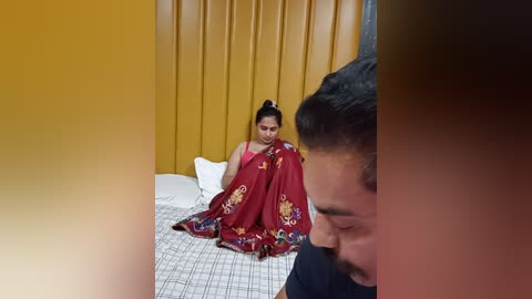 Media: Video of a South Asian woman in a red sari with floral embroidery, seated on a bed with white sheets, in a room with mustard-yellow curtains. A man, partially visible, is seen in the foreground.