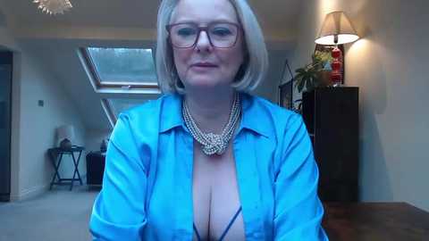 Media: Video of a fair-skinned woman with short blonde hair, glasses, and a blue blouse revealing cleavage. She sits in a modern living room with beige walls, a lamp, and a plant.