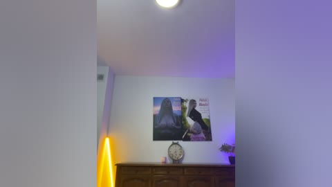 Media: Video of a minimalist living room with white walls, a wooden dresser, a large framed photo of two people, a tall yellow lamp, and a wall clock.