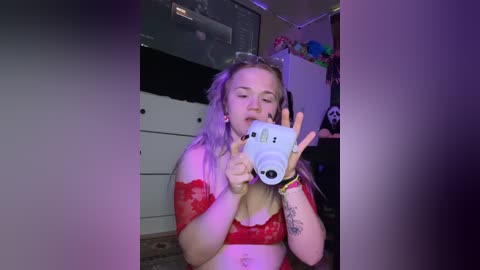 Media: Video of a young woman with long, wavy blonde hair, wearing red lace lingerie, taking a selfie with a white camera. She has a tattoo on her right forearm. Background shows a dimly lit room with a white dresser, colorful plush toys, and a black wall.