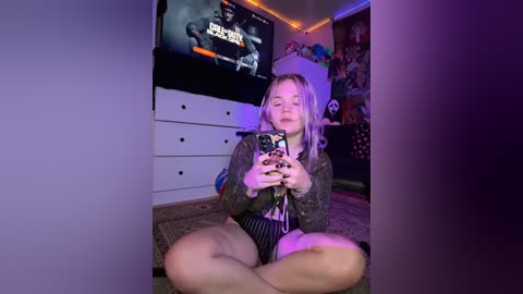 Media: A video of a young woman with long blonde hair, sitting cross-legged on a rug, playing a video game in a dimly lit room with a TV, dresser, and posters.