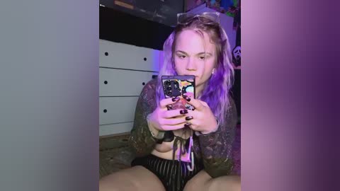 Media: Video of a young woman with pink hair, wearing a cropped jacket, sitting on the floor, taking a selfie in a dimly lit room with purple lighting.