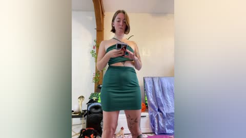 Media: Video of a woman with short blonde hair, in a green strapless crop top and high-waisted skirt, standing in a cluttered room with a wooden archway, plants, and a blue tarp in the background.