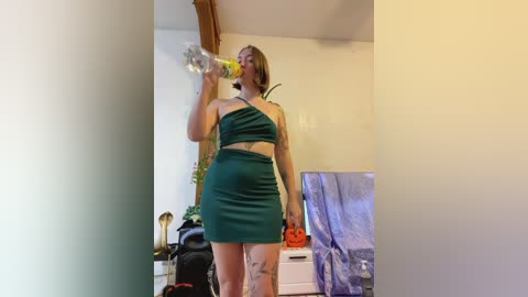 Media: Video of a plus-sized, light-skinned woman with short brown hair, wearing a green halter top and matching mini skirt, drinking water in a cluttered, dimly lit bedroom with a white dresser and TV.