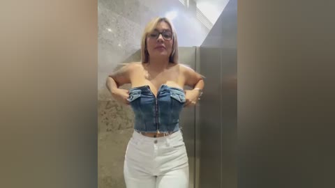 Media: Video of a blonde woman with glasses in a strapless denim crop top and high-waisted white pants, standing in a dimly lit, beige-tiled hallway.