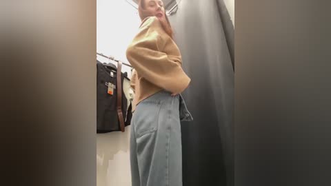 Media: A video captures a woman in a dressing room, wearing a tan sweater and high-waisted light blue jeans, standing in front of a black garment on a hanger.