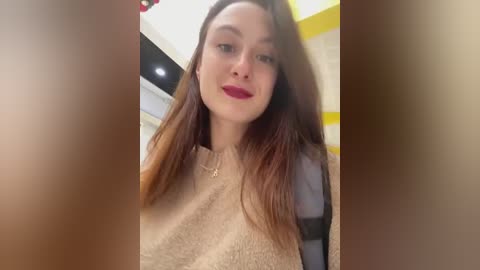 Media: Video of a fair-skinned woman with long, straight brown hair, wearing a beige sweater and red lipstick, standing indoors with a yellow and white background.