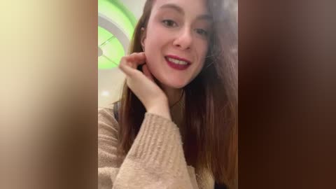 Media: Video of a young woman with long brown hair and fair skin, smiling, with green neon light background, wearing a beige knit sweater.