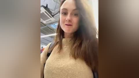 Media: A video of a young Caucasian woman with light skin and long brown hair, wearing a beige sweater, standing in an airport with white ceiling panels and visible luggage.