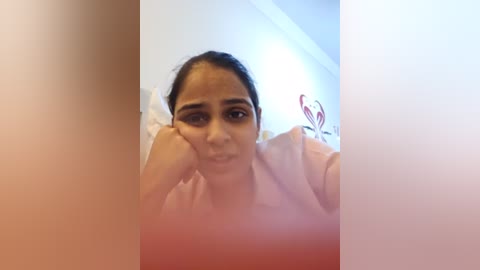 Media: A video of a South Asian woman with dark hair and medium skin tone, wearing a light pink top, resting her head on her hand, smiling slightly. Background includes a white wall with red heart stickers.