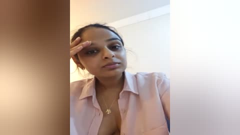 Media: A video of a young South Asian woman with medium brown skin, dark hair tied back, wearing a light pink blouse, touching her forehead. She has a small gold necklace and neutral expression. Background is a white wall.