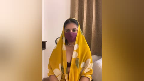 Media: A video of a South Asian woman with medium skin tone and dark hair, wearing a yellow sari with white patterns, a purple mask, and a pearl necklace. She sits in a beige room with a wooden headboard in the background.