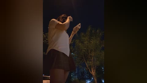 Media: A video of a woman wearing a white top and black skirt, holding a phone, at night against a dark background with trees.