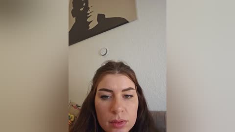 Media: A video of a young woman with long brown hair and dark eyebrows, wearing makeup, standing in a room with a cream-colored wall featuring a black silhouette of a person, and a small clock.