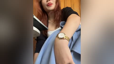 Media: A video of a young woman with fair skin and long, reddish-brown hair, wearing a black cardigan over a white top, red lipstick, and a gold watch. She is holding a blue shirt, in a dimly lit room.