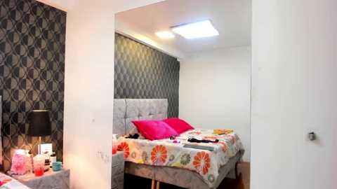 Media: Video of a modern, minimalistic bedroom with a bed dressed in floral sheets, bright pillows, a patterned wall, and a small nightstand with a lamp.