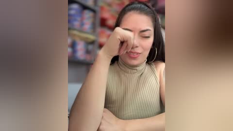 Media: Video of a young Latina woman with medium skin tone, long dark hair, wearing a beige ribbed turtleneck sweater. She has her right hand on her forehead, looking tired. Background shows colorful snack aisle.