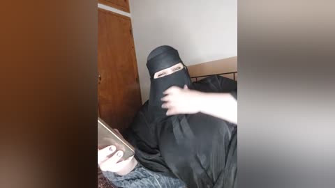 Media: Video of a person in a black niqab, sitting on a bed in a dimly lit room with a wooden door in the background.