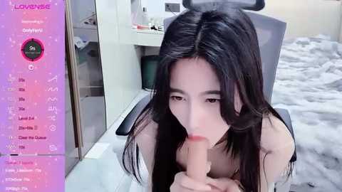 Media: Video of an East Asian woman with long black hair, performing oral sex on a realistic, flesh-colored dildo, in a bedroom setting with a gray carpet and white furniture.