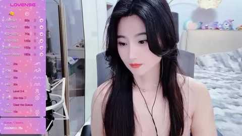 Media: Video of a young Asian woman with long black hair, wearing a sleeveless top, sitting in a modern bedroom with a pink background and a gray bed.