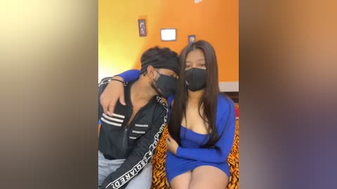 Media: Video of two Asian individuals in a room with orange walls; one wearing a black jacket and mask, the other in a blue dress and mask, sitting close together on a tiger-print blanket.