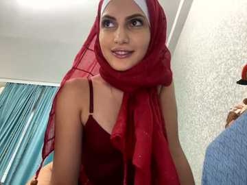 Media: Video of a young woman with light skin and dark eyes, wearing a red hijab and maroon dress, smiling slightly, indoors with light blue curtains and textured wallpaper in the background.