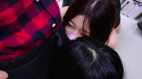 Media: Video of a woman with long black hair, wearing a red and black plaid shirt, kissing a man with black hair. They are in a clinical setting with medical equipment visible in the background.