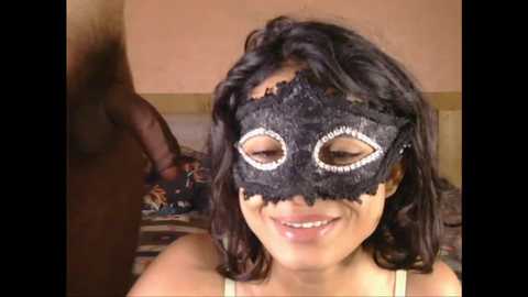 Media: Video of a smiling young woman with medium skin tone wearing a black lace masquerade mask with white sequins, seated indoors, with a dark-skinned man partially visible in the background.