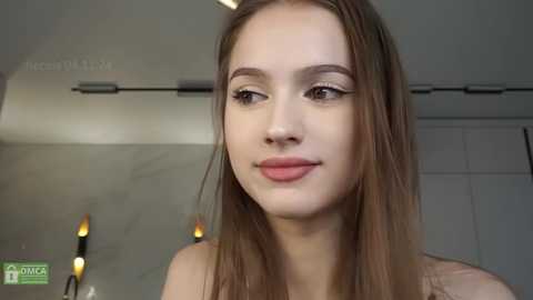 Media: A video of a young Caucasian woman with straight, light brown hair, light skin, and subtle makeup, smiling slightly. She wears a sleeveless top. Background includes a grey wall and a door.