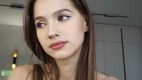 Media: Video of a young Caucasian woman with long brown hair, light skin, and natural makeup, including subtle eyeliner. She has a neutral expression. Background features a modern, minimalist interior with white walls and a closet.