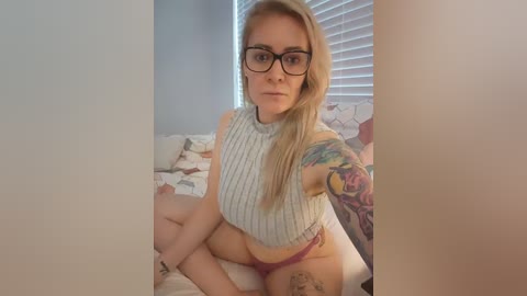 Media: Video of a young woman with long blonde hair, glasses, and a sleeve tattoo, wearing a ribbed crop top and pink panties, sitting on a bed with floral bedding.