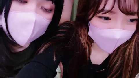 Media: Video of two Asian women with light skin, wearing white face masks, dark hair, and black clothes, looking serious, taken indoors with a blurred background.