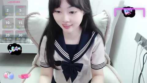 Media: A video of an East Asian woman with long black hair, fair skin, and a petite frame, wearing a Japanese schoolgirl uniform, sitting in a modern living room.