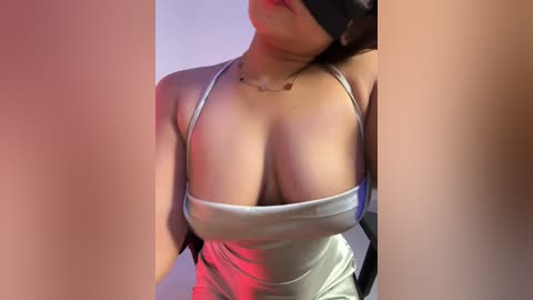 Media: Video of a woman with fair skin, wearing a black blindfold, a silver tank top revealing ample cleavage, and a necklace. She is indoors with a blurred background.