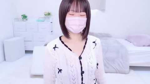 Media: Video of an Asian woman with straight black hair, wearing a white blouse with black bow patterns and a pink face mask, standing in a bright, minimalist bedroom with white furniture and light-colored bedding.