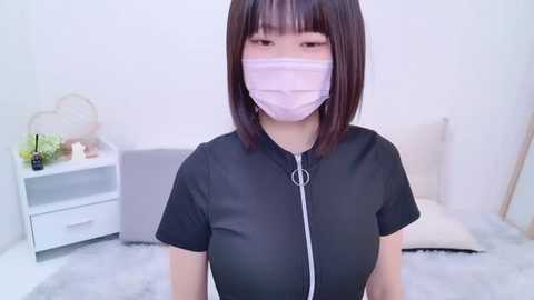 Media: Video of an Asian woman with straight, shoulder-length black hair, wearing a black zip-up top and a pink face mask, standing in a white bedroom with a white bed, white nightstand, and a plush gray rug.