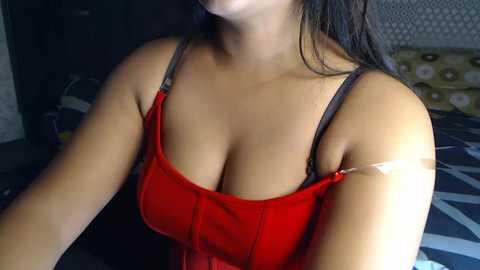 Media: Video of a young woman with light brown skin, wearing a low-cut red velvet dress with thin straps, revealing ample cleavage. She has long, dark hair. Background includes a patterned wall and a bed with white pillows.