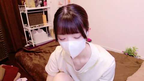 Media: Video of a young Asian woman with straight black hair and bangs, wearing a white mask, sitting on a brown carpet in a living room with a white metal shelving unit and a brown chair in the background.