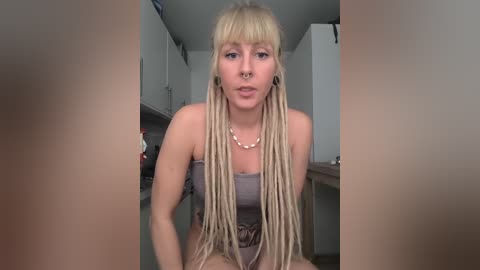 Media: Video of a light-skinned woman with long, blonde dreadlocks, wearing a brown strapless top, standing in a modern kitchen with white cabinets. She has a septum piercing and black ear gauges.