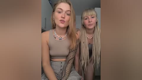 Media: Video of two young women with fair skin and long, blonde hair. One wears a beige crop top, high-waisted patterned pants, and a beaded necklace. The other has dreads, a black crop top, and a similar necklace. They sit side by side, indoors, with blurred background.