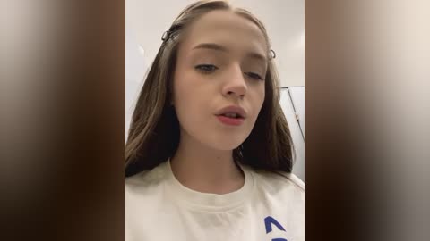 Media: A video of a young Caucasian woman with long brown hair, wearing a white shirt, captured from a slightly blurry, close-up angle.