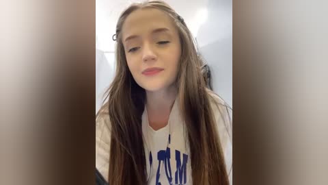 Media: Video of a young girl with long, straight brown hair, wearing a white t-shirt with blue text. She has fair skin and a calm expression, standing indoors with blurred background.