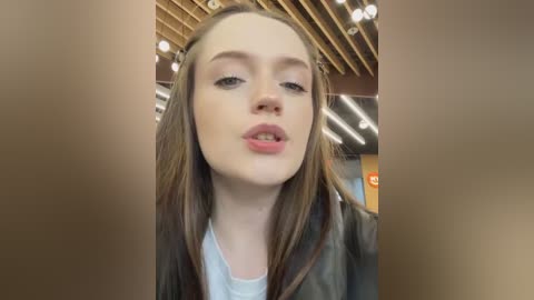 Media: Video of a young Caucasian woman with fair skin, brown hair, and light makeup, wearing a black jacket and white top. She's indoors, with a wooden ceiling and fluorescent lighting in the background.