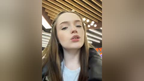 Media: A close-up video of a young woman with long, straight brown hair, wearing a black jacket over a white shirt. She has a light complexion and is blowing a kiss. The background features a modern, wooden ceiling with LED lights.