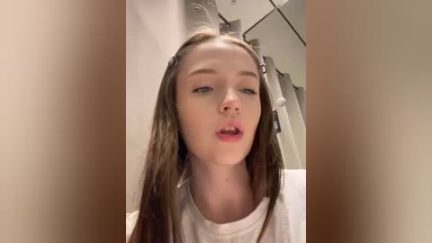 Media: Video of a young Caucasian woman with fair skin, long brown hair, wearing a white t-shirt, looking expressionless in a bathroom setting.