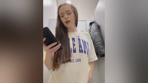 Media: Video of a young Caucasian woman with long brown hair, wearing a white t-shirt with blue text, holding a phone. She stands in a dimly lit hallway, with a black jacket hanging nearby.