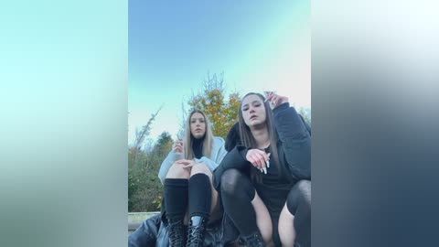 Media: Video of two young women, one with long hair, wearing a hoodie, and the other with a black top and jacket, sitting on a park bench under a clear blue sky with autumn trees in the background.